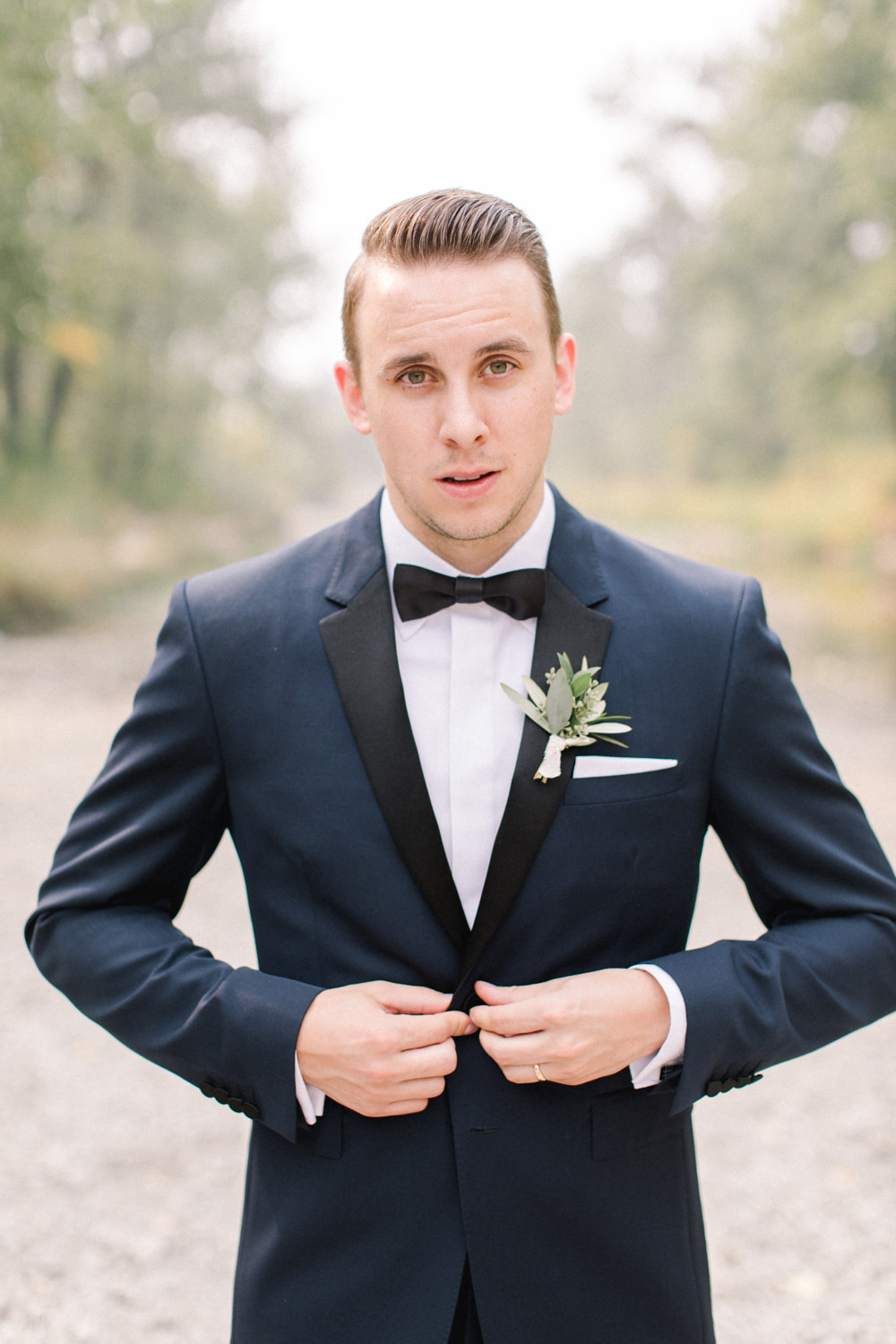 portrait of groom buttoning jacket at fish creek park in Calgary at the lake house in Calgary