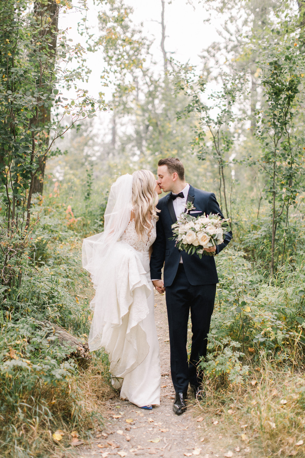wide shot of the bride and groom walking while sharing a kiss in fish creek park in Calgary at the lake house in Calgary