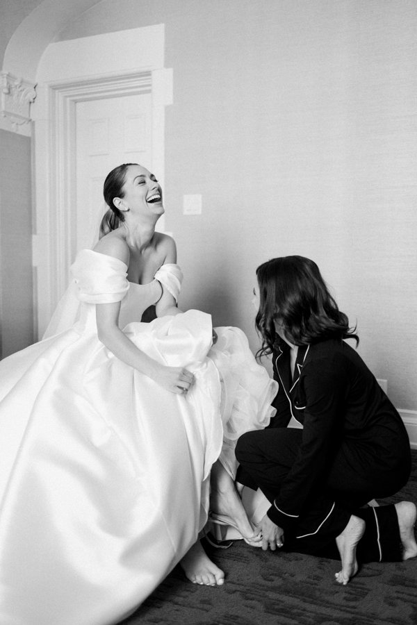 black and white photo of bride getting help putting on her wedding day shoes laughing with sister at the Calgary Fairmont Palliser Hotel